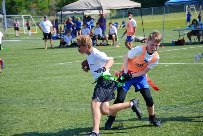 player in a white shirt and wearing sunglasses runs with a football in his arm while a player in an orange jersey pulls his flag